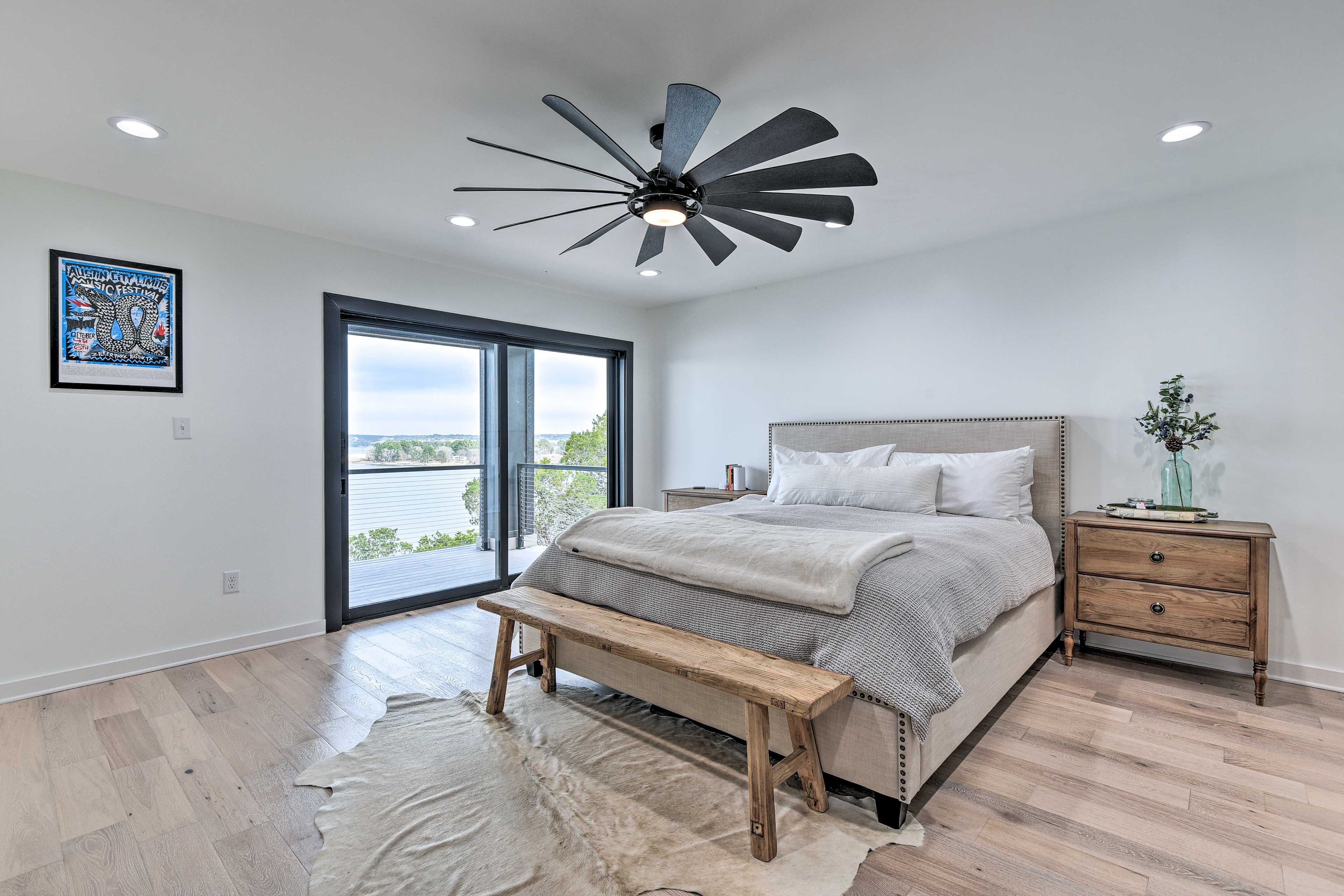 A bedroom with a large bed, a bench, a nightstand, a ceiling fan, and a window with a view.