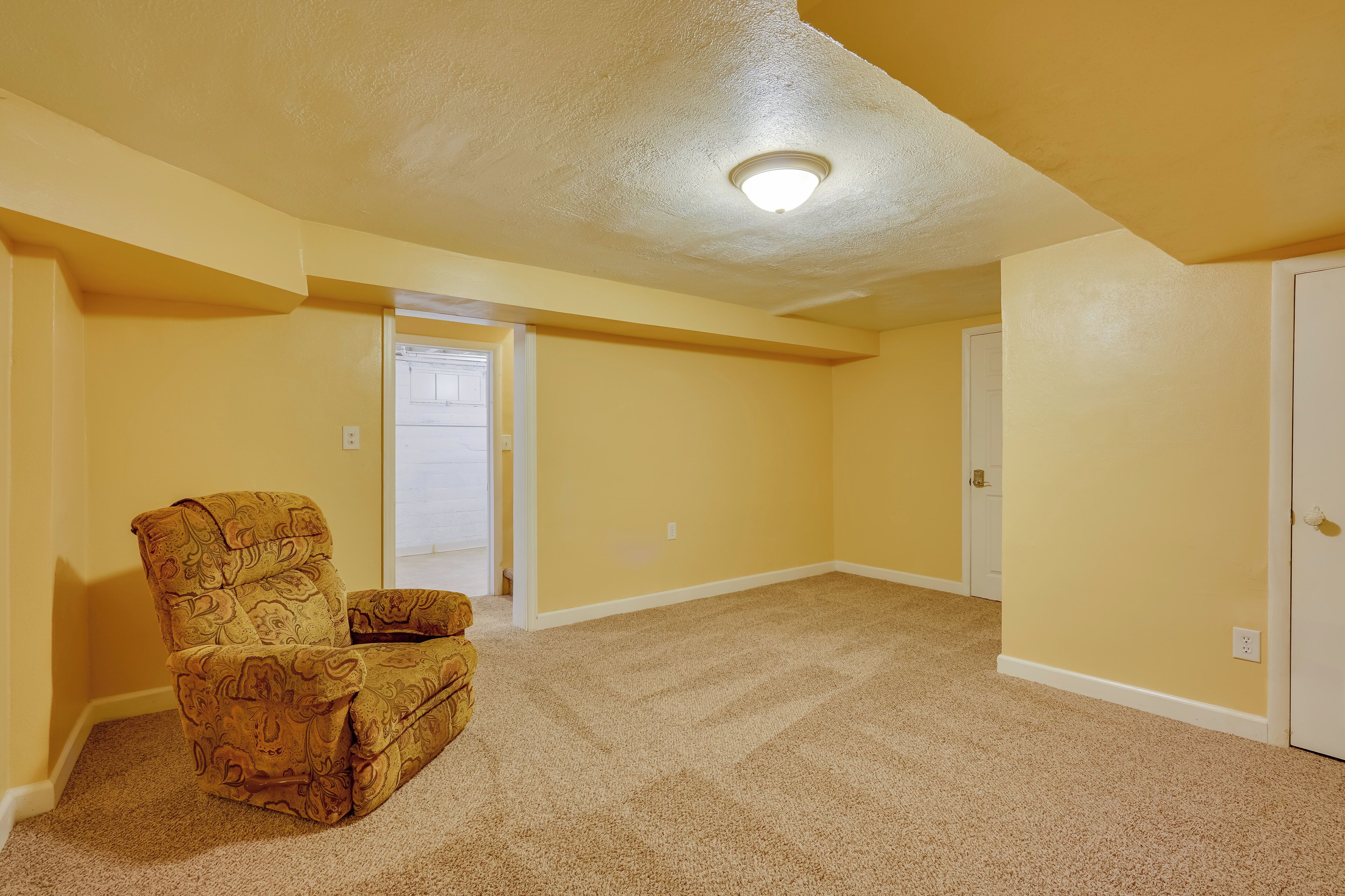 Una habitación con paredes amarillas, piso alfombrado, un sillón estampado y una puerta que da a otra habitación.
