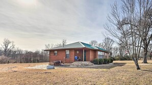 A single-story brick building with a green roof, a front porch, and a small yard. - Pet-friendly Cabin Rental Retreat! (Keota)