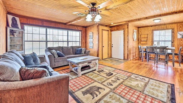 A spacious living room with a wooden ceiling, a ceiling fan, a fireplace, a dining area with a table and chairs, and a large window providing natural light. - Pet-friendly Cabin Rental Retreat! (Keota)