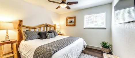 Un dormitorio con cabecera de madera, cama con colcha blanca y gris, ventilador de techo, mesita de noche con lámpara, un cuadro en la pared y ventana con persianas.