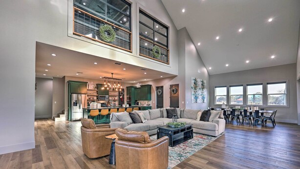 A spacious living area with a large sectional sofa, leather armchairs, and a dining table with chairs. A kitchen is visible in the background, and there are large windows providing natural light. - Lavish Riverfront Escape w/ Gas Grill! (Glenwood)