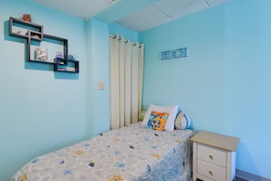 A bedroom with a bed, a nightstand, a shelf with decorative items, and a curtain. - Simple North Wildwood Condo - Steps to Beach! (North Wildwood)