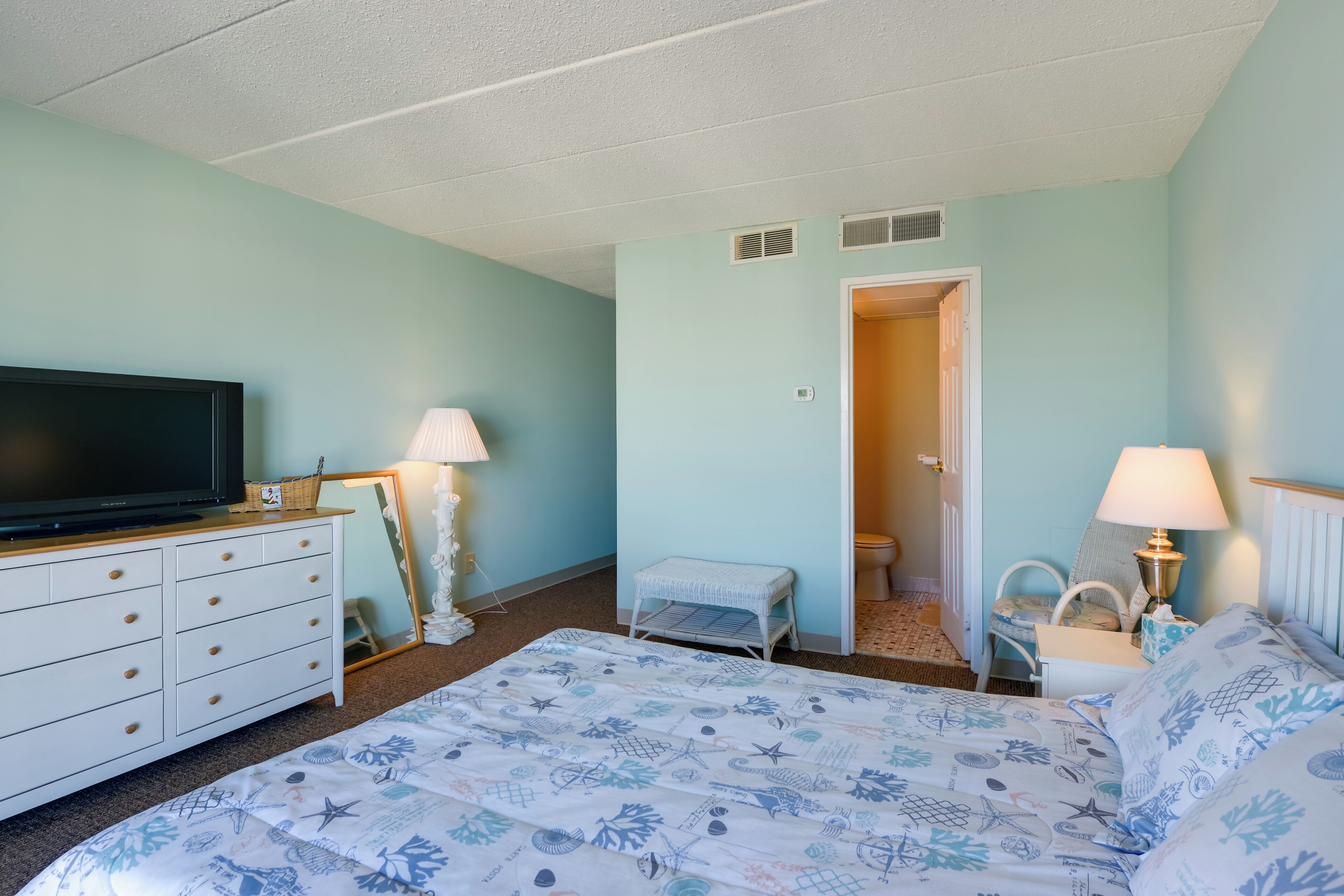A bedroom with a bed, a dresser, a television, a lamp, and a door leading to a bathroom.
