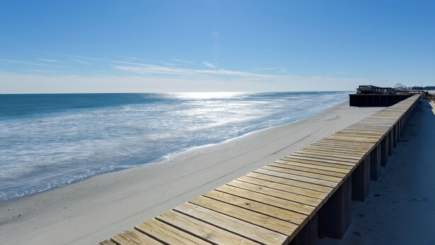 A wooden boardwalk along a beach with a clear sky and ocean in the background.