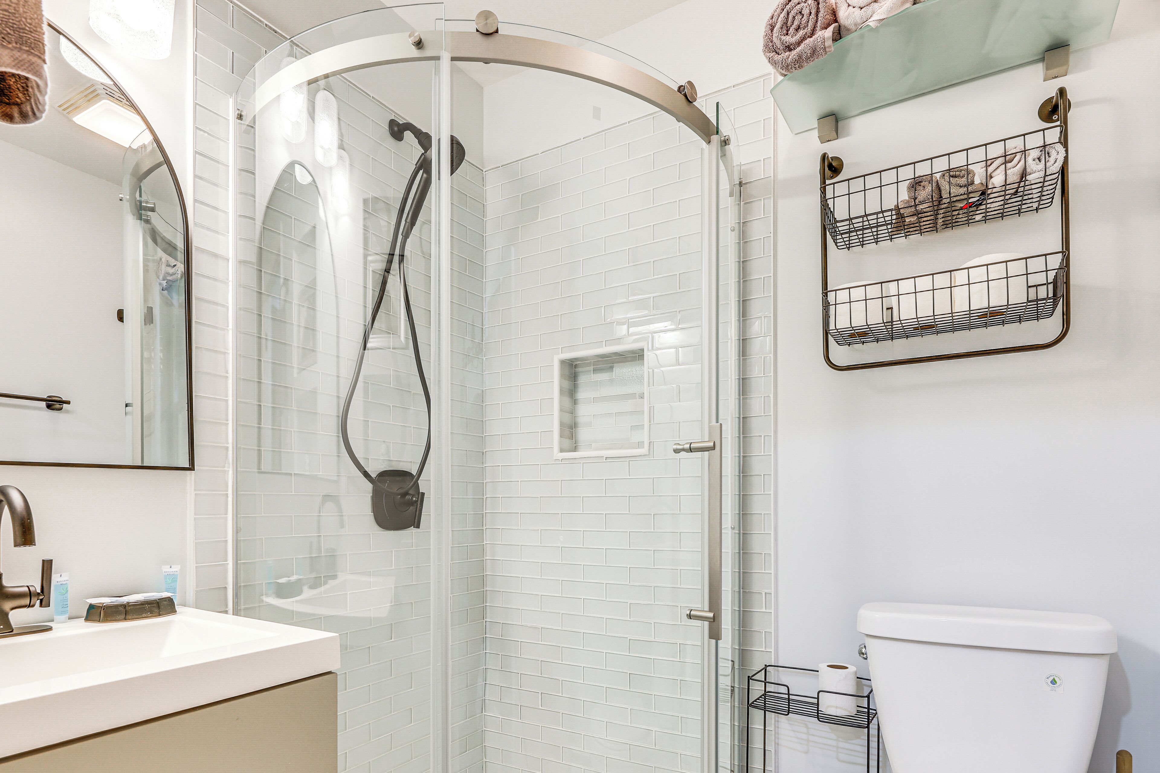 A modern bathroom with a glass-enclosed shower, a white-tiled wall, a sink with a mirror, and a toilet with a storage basket.