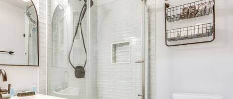 A modern bathroom with a glass-enclosed shower, a white-tiled wall, a sink with a mirror, and a toilet with a storage basket.
