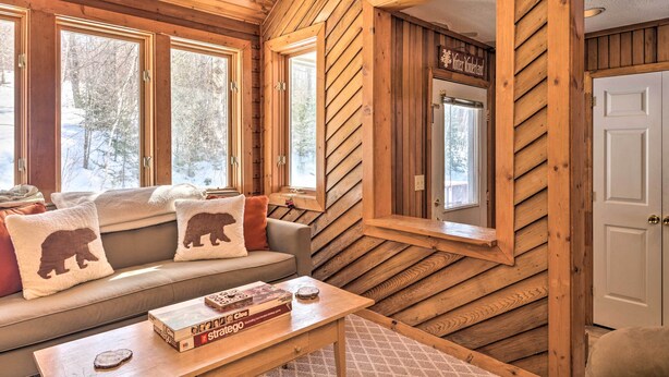 A cozy cabin interior with a wooden interior, a sofa with bear-themed pillows, a coffee table with books, and a mirror reflecting the door. - Updated Condo Next to Burke Mountain Bike Trails! (East Burke)