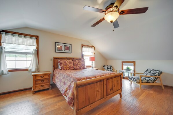 A bedroom with a wooden bed, a ceiling fan, a nightstand, a chair, and a window with curtains. - Quaint Fredericksburg Studio w/ Shared Hot Tub (Fredericksburg)