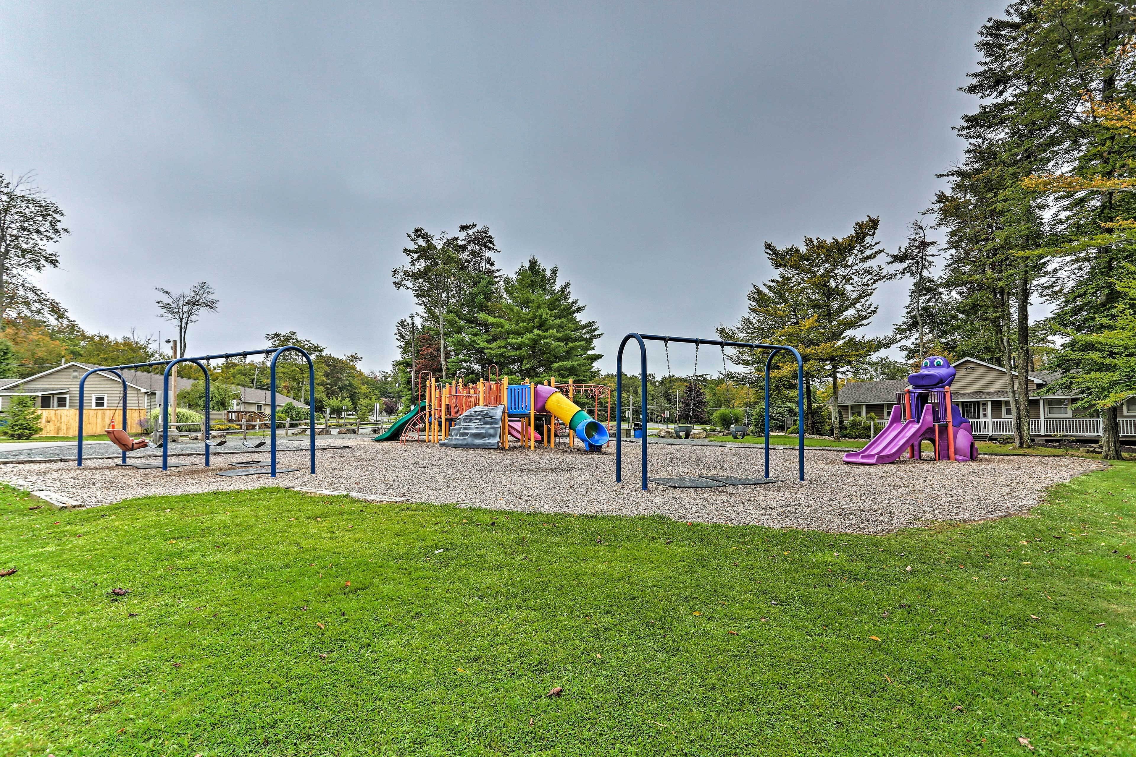 A playground with various play structures including slides and swings, surrounded by a grassy area and trees.