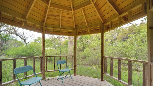 Un gazebo in legno con tetto, due sedie blu e un patio in legno con vista su una zona boschiva.