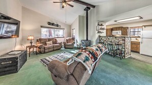 A cozy living room with a fireplace, a ceiling fan, and a kitchen area with wooden cabinets and a refrigerator.