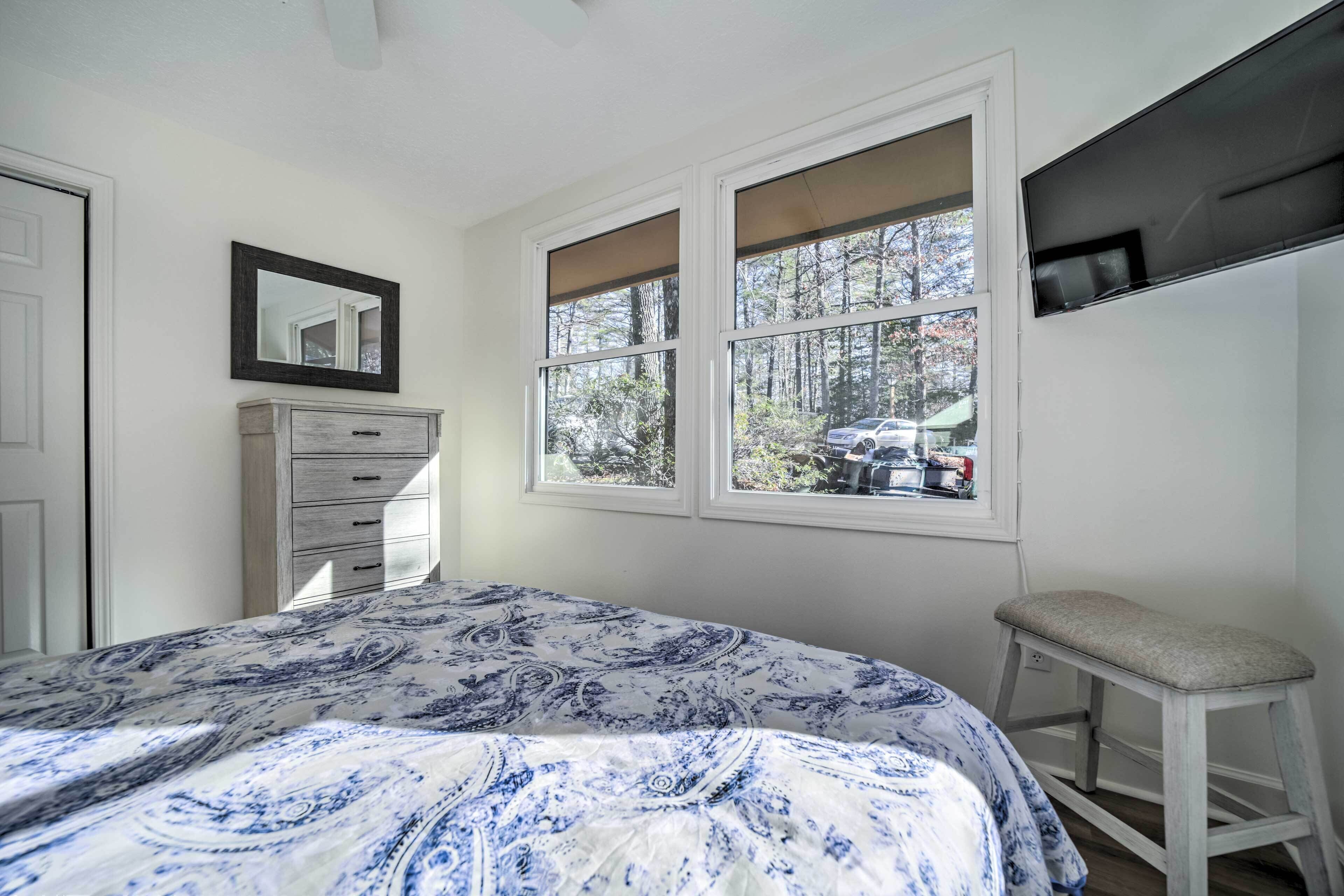 A bedroom with a bed, a dresser, a window with a view of trees and a car, and a TV mounted on the wall.