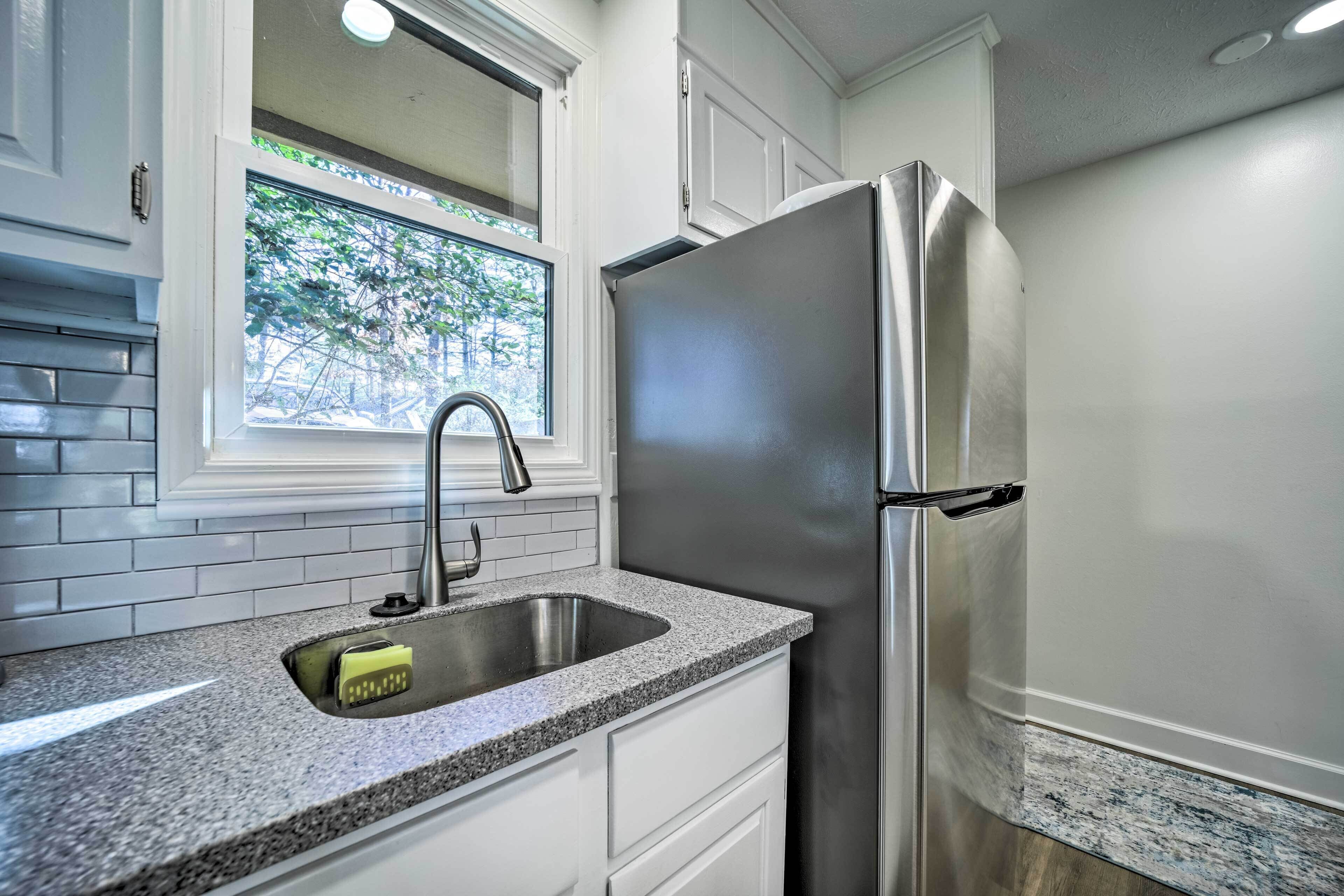 Una cocina con armarios blancos, un refrigerador de acero inoxidable, una encimera de granito y una ventana con vistas a árboles.