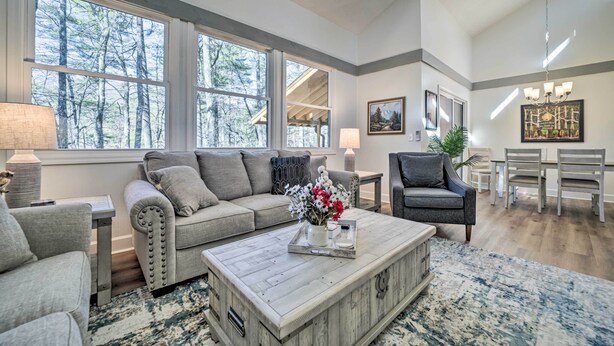 A spacious living room with a large window, a grey sofa, a coffee table, and a dining area with chairs. - Sapphire Home w/ Grill & Pool Access (Sapphire)