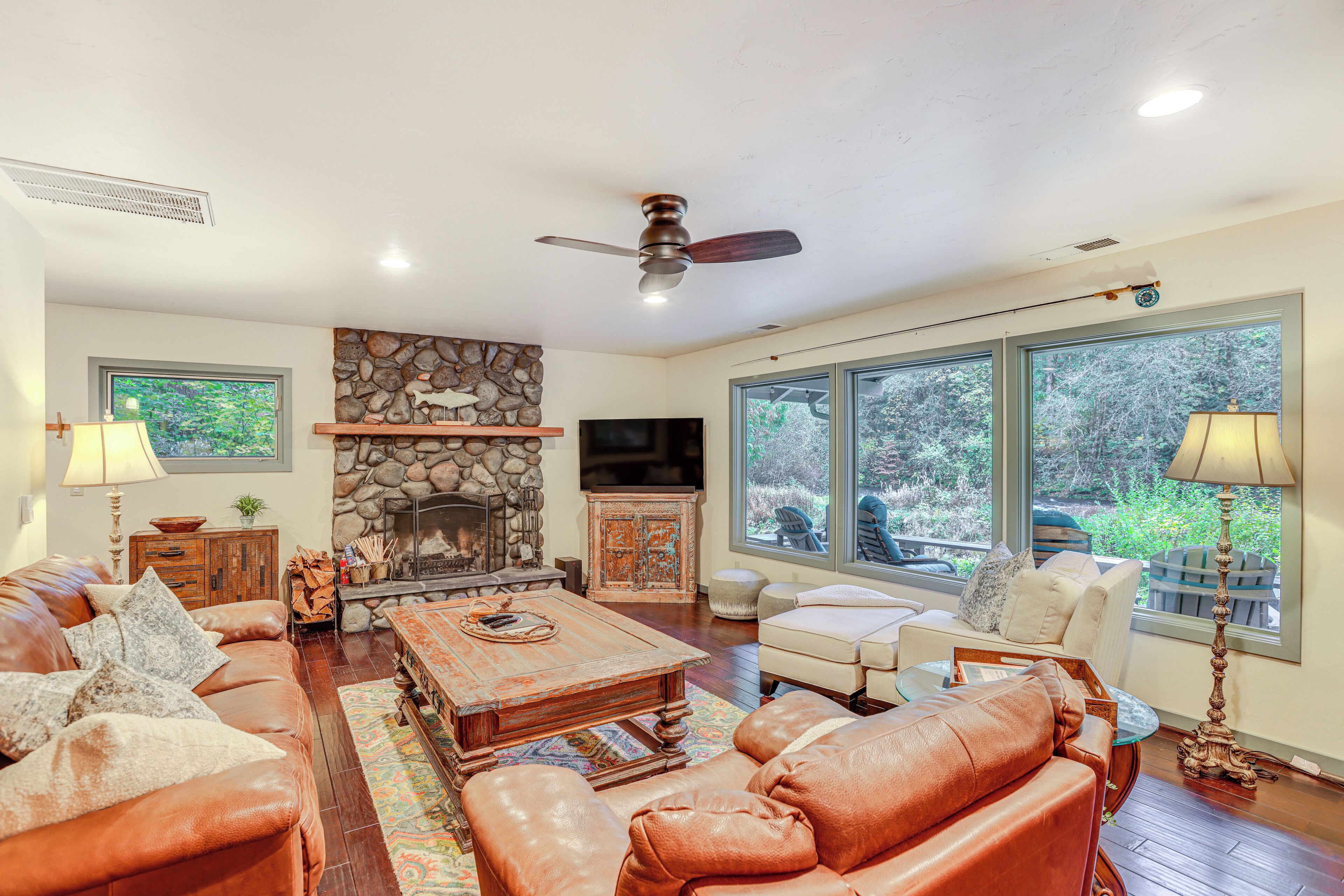 A spacious living room with a stone fireplace, leather sofas, a coffee table, and large windows offering a view of the outdoors.