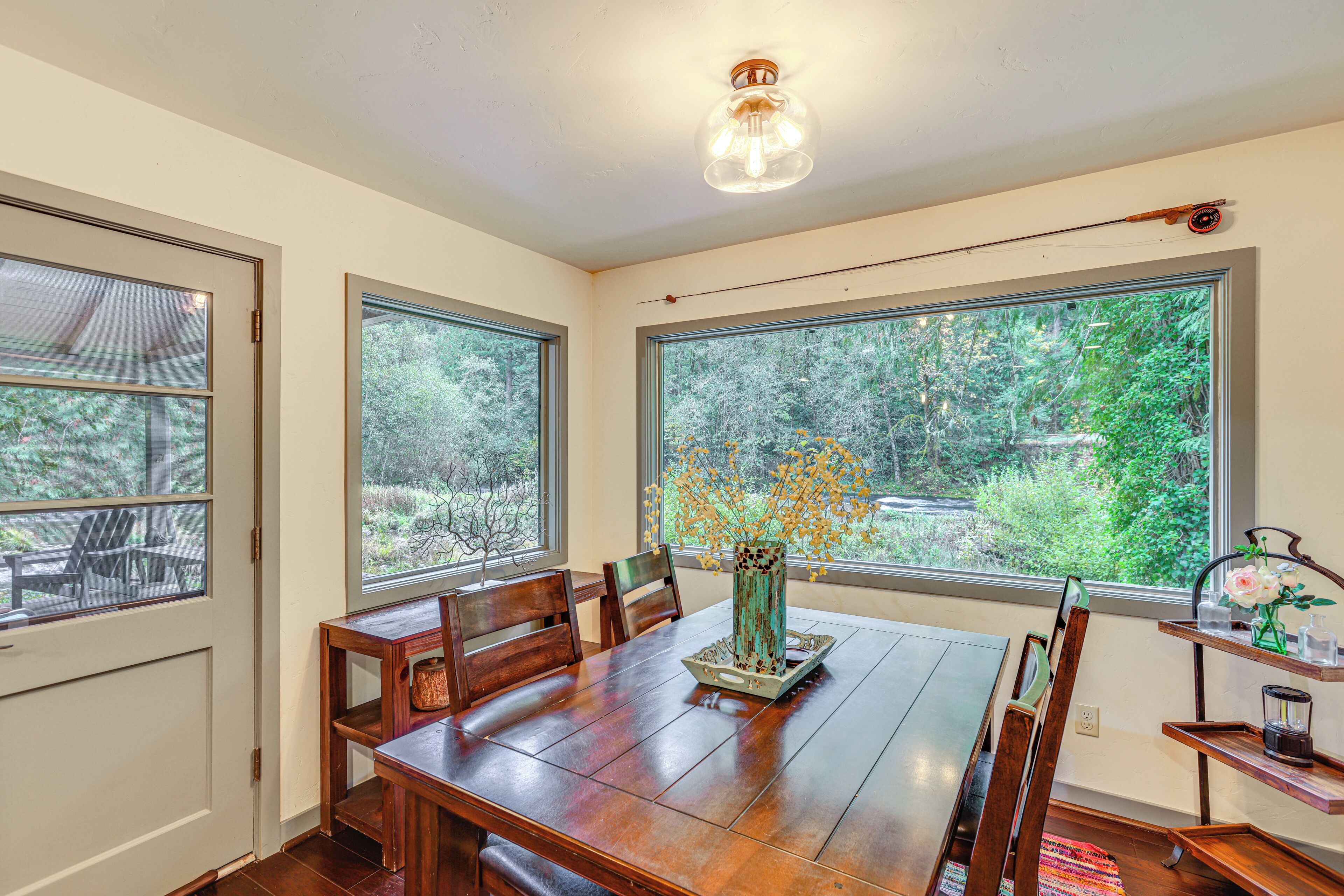 A dining room with a wooden table, chairs, and a vase with flowers. There are large windows with a view of greenery outside.