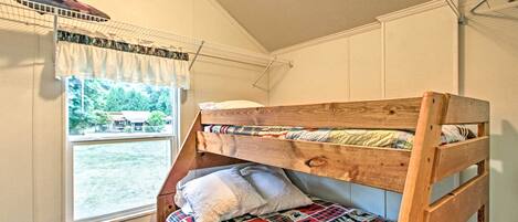 A bunk bed with a patterned quilt, a small bedside table, and a window with a view of a house and trees.