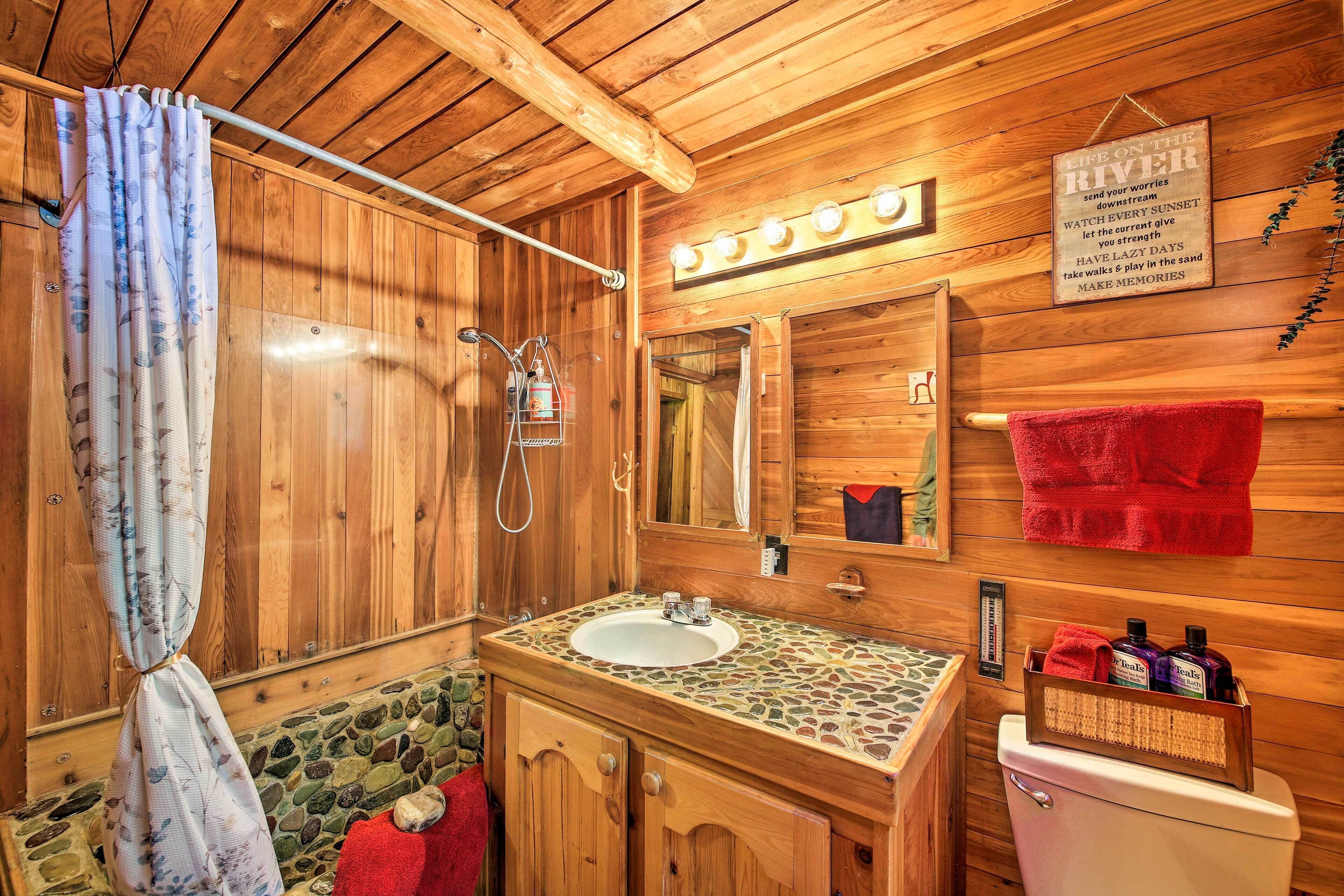 A wooden cabin bathroom with a stone sink, a mirror, a shower, and a sign that reads "Life on the River."