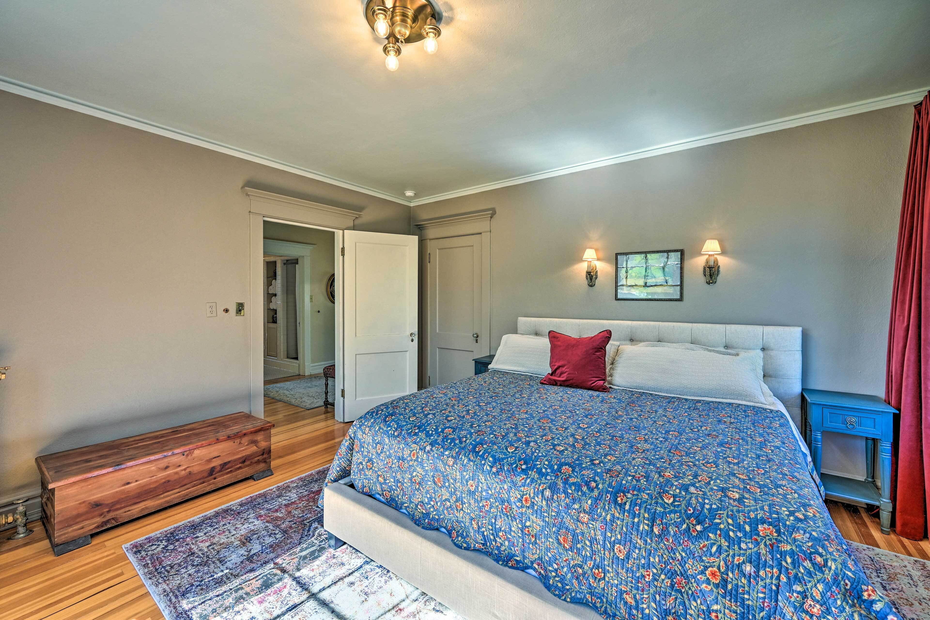 A bedroom with a large bed, a wooden bench, a nightstand, and a wall-mounted lamp.