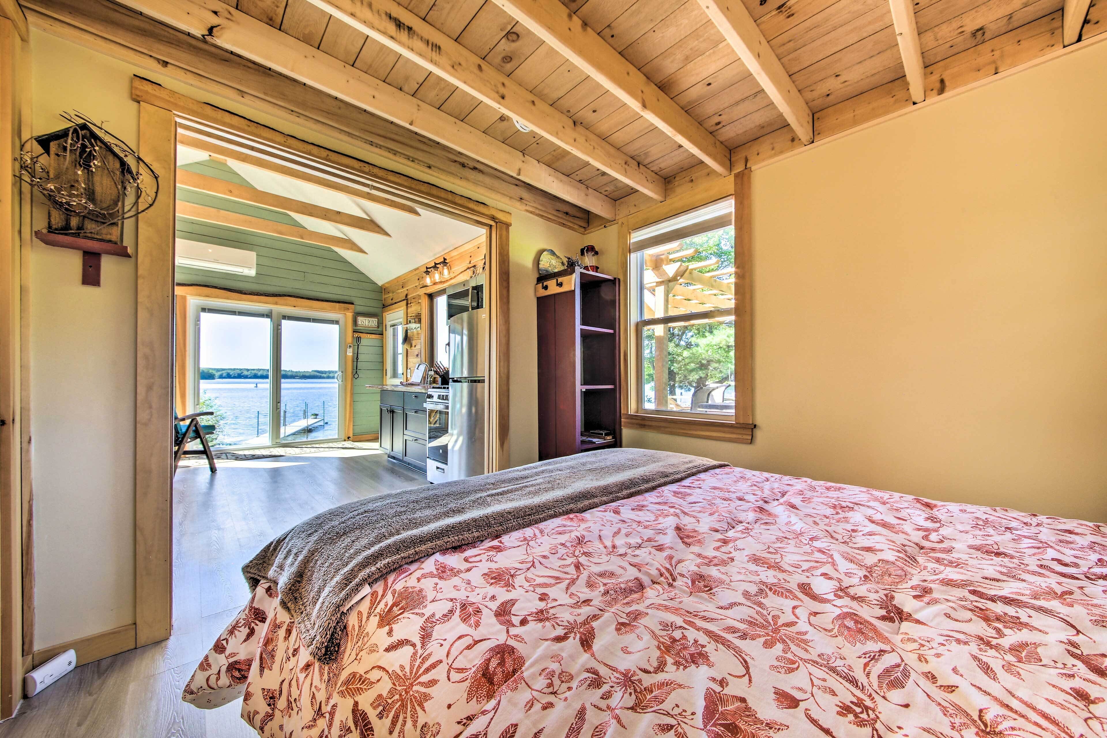 A bedroom with a large bed, wooden ceiling beams, a view of the outdoors, and a kitchenette area.