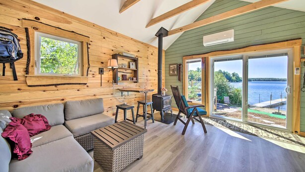 A cozy cabin interior with a sofa, a table, and a chair. There is a window with a view of the outdoors, a fireplace, and a door leading outside. - Quaint Oakland Getaway on East Pond Lake! (Oakland)