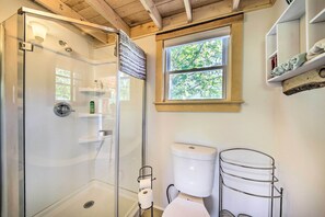 Ein Badezimmer mit einer gläsernen Duschkabine, einer Toilette mit einem Metallregal und einem Fenster mit Blick auf Bäume.