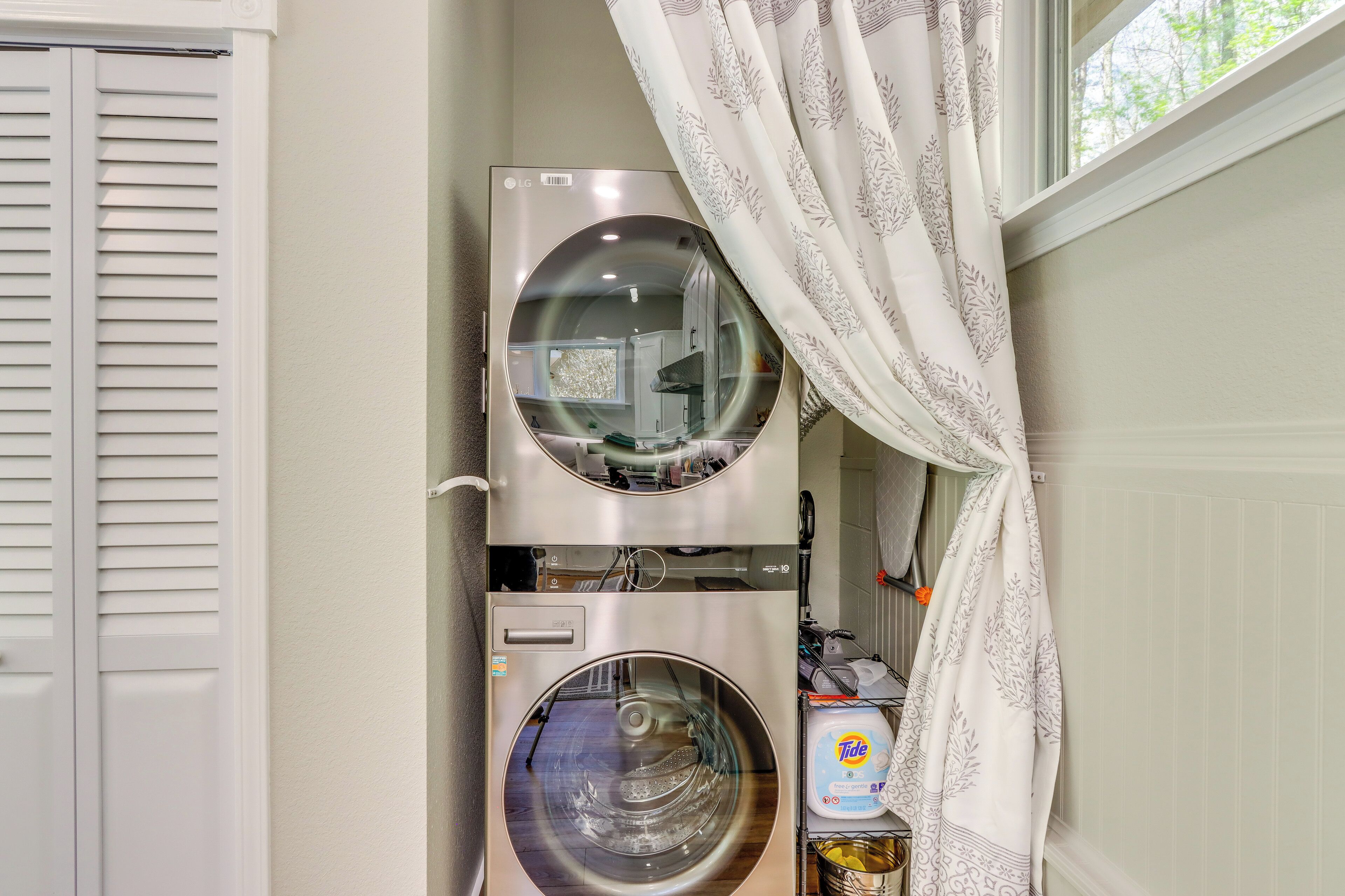 Une petite buanderie comprenant deux machines à laver et deux sèche-linge superposés, une étagère avec des produits de lessive et une fenêtre avec des rideaux.