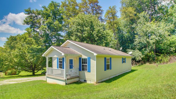 A single-story house with a porch, surrounded by lush greenery and trees. - Walk to Main St: Inviting Byrdstown Home w/ Yard! (Byrdstown)
