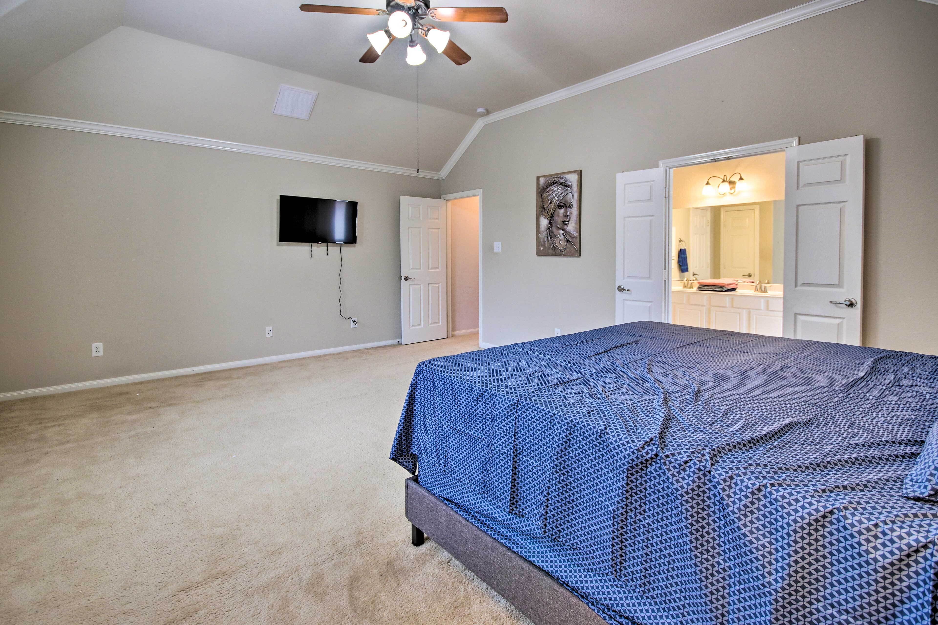 A bedroom with a large bed, a ceiling fan, a TV mounted on the wall, and a bathroom with a mirror and sink.