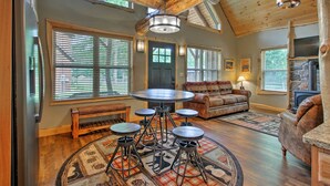 A rustic interior with a wooden ceiling, a stone fireplace, a round dining table with stools, a sofa, and a large window with blinds. - Timber Home: Walk to Slopes & N Conway Shops! (North Conway)