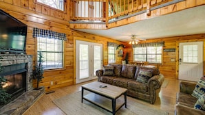 A cozy cabin interior with a fireplace, wooden walls, a loft bedroom, and a leather sofa. - Sevierville Cabin w/ Hot Tub, Views & Pool Access! (Sevierville)