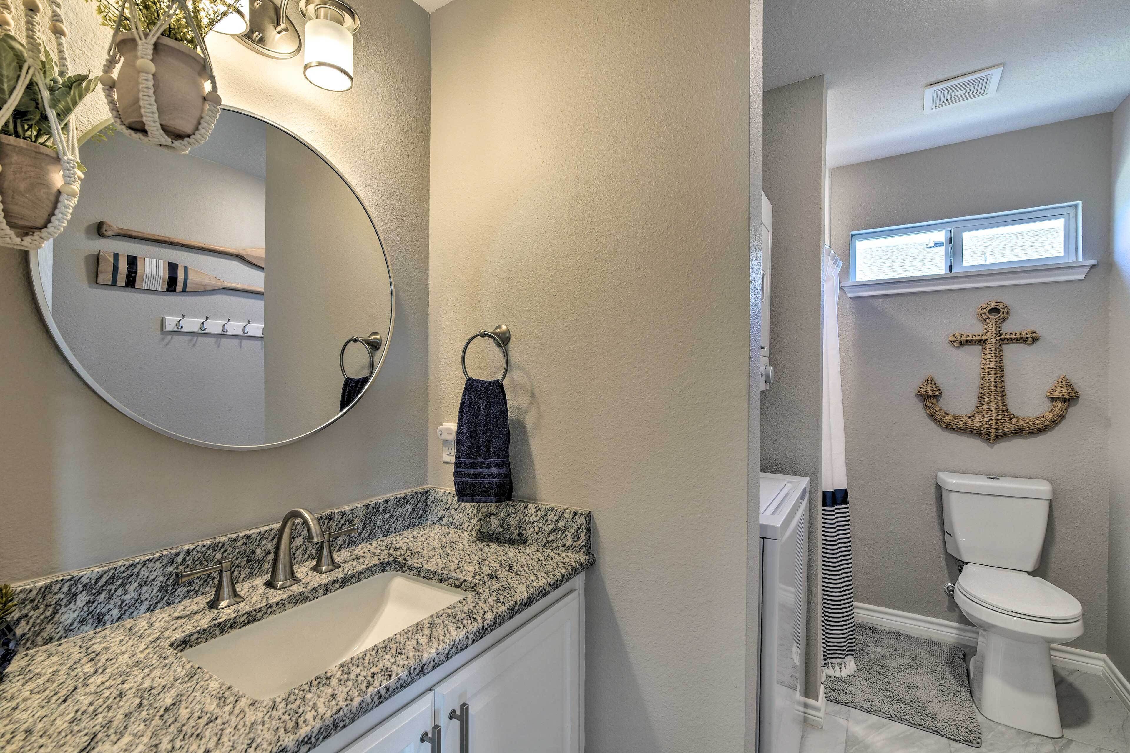 A bathroom with a granite countertop sink, a round mirror, and a toilet with a decorative anchor wall hanging.