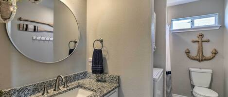 A bathroom with a granite countertop sink, a round mirror, and a toilet with a decorative anchor wall hanging.