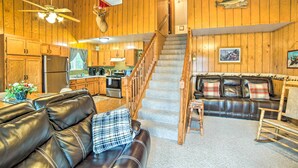 A spacious living area with a leather sofa, a staircase, wooden walls, a kitchen, and a dining area. - Secluded Woodsy Watersmeet Cabin w/ Fire Pit! (Watersmeet)