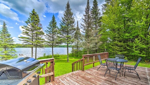 A wooden deck with a grill, a table with chairs, and a view of a lake surrounded by trees. - Secluded Woodsy Watersmeet Cabin w/ Fire Pit! (Watersmeet)