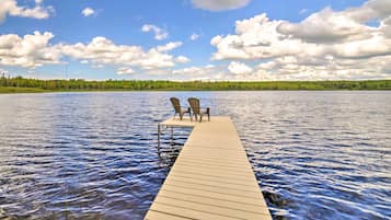 A wooden dock extending into a calm lake with two chairs facing the water, surrounded by a forested landscape under a partly cloudy sky.