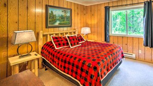 A bedroom with wooden walls, a bed with a red and black checkered bedspread, two bedside lamps, a painting of a landscape, and a window with curtains. - Secluded Woodsy Watersmeet Cabin w/ Fire Pit! (Watersmeet)