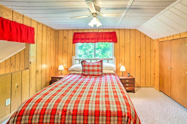 A bedroom with wooden paneling, a plaid bedspread, a window with a red valance, a ceiling fan, and a nightstand with a lamp.