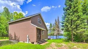 A brown single-story cabin with a front door and a small porch, surrounded by a fenced yard and trees, near a body of water. - Secluded Woodsy Watersmeet Cabin w/ Fire Pit! (Watersmeet)