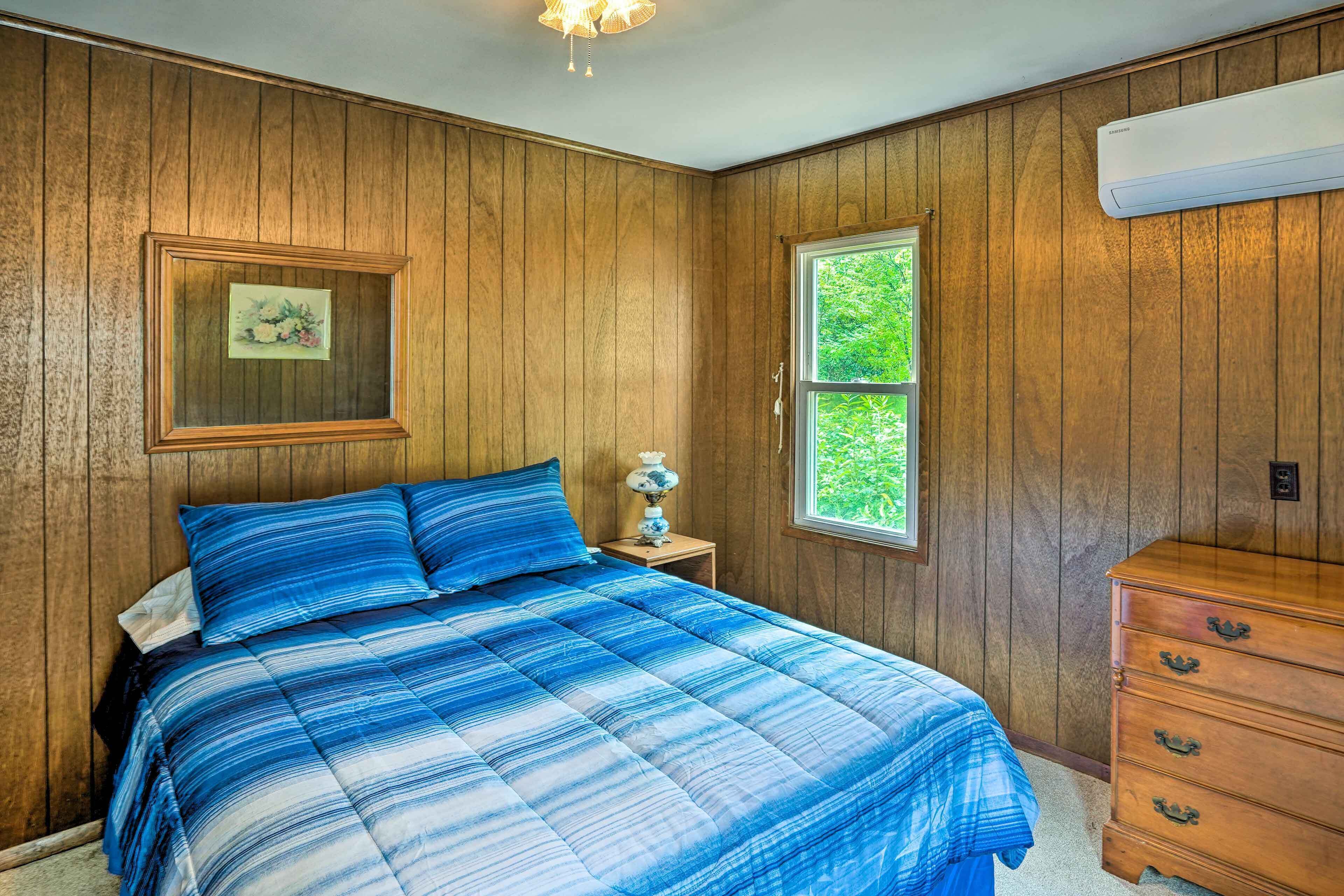 A bedroom with wooden paneling, a bed with blue bedding, a nightstand with a lamp, a dresser, and a window with a view of greenery.