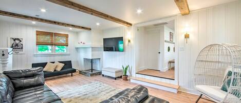 Un salón espacioso con un sofá de cuero negro, un sillón blanco y un banco acolchado blanco. Un televisor de pantalla plana está montado en la pared, y hay una escalera de madera que conduce a otra habitación.