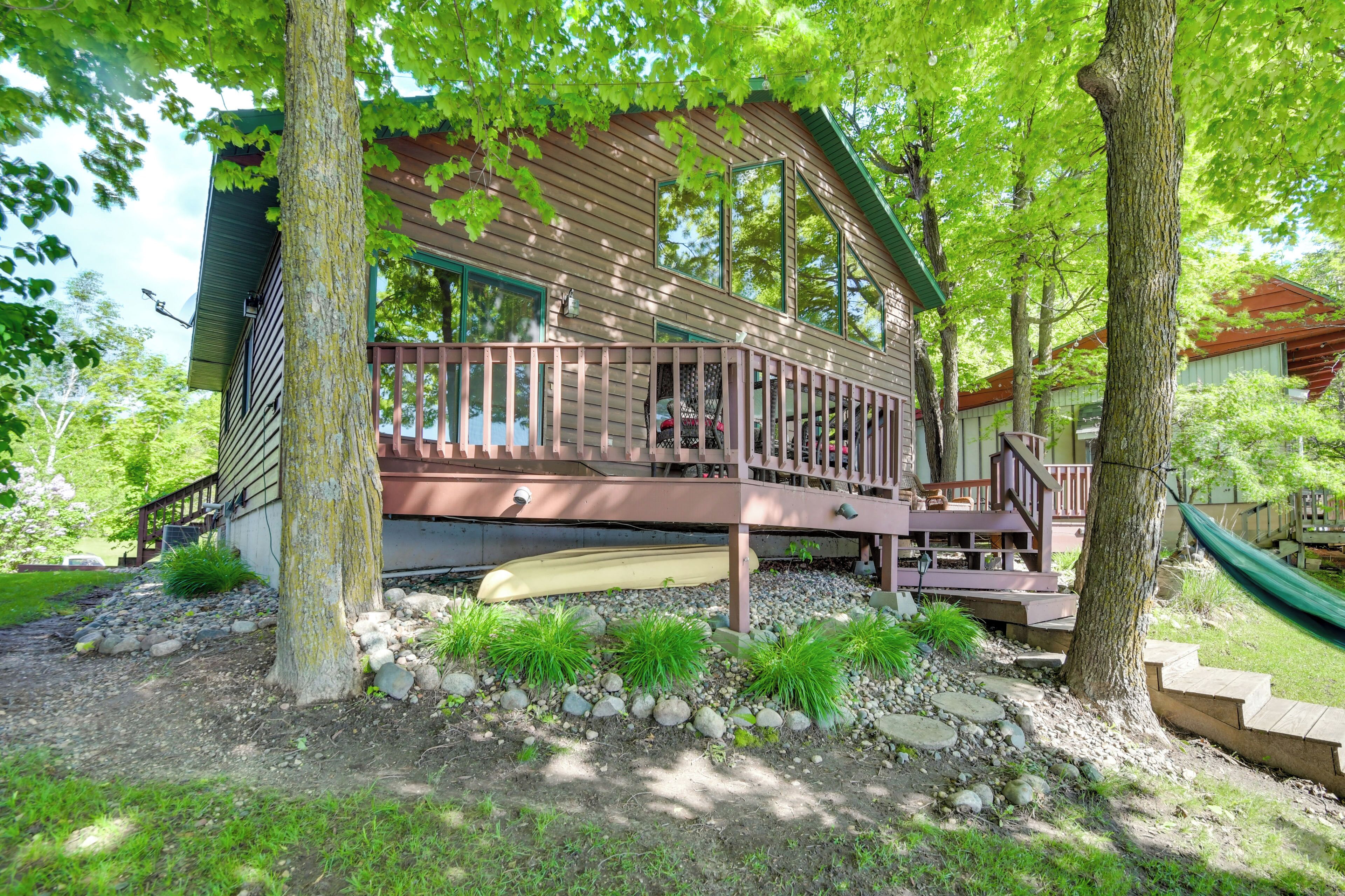 A wooden cabin with a deck, surrounded by trees and a gravel path.
