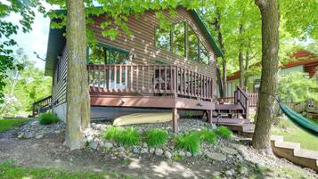 A wooden cabin with a deck, surrounded by trees and a gravel path.