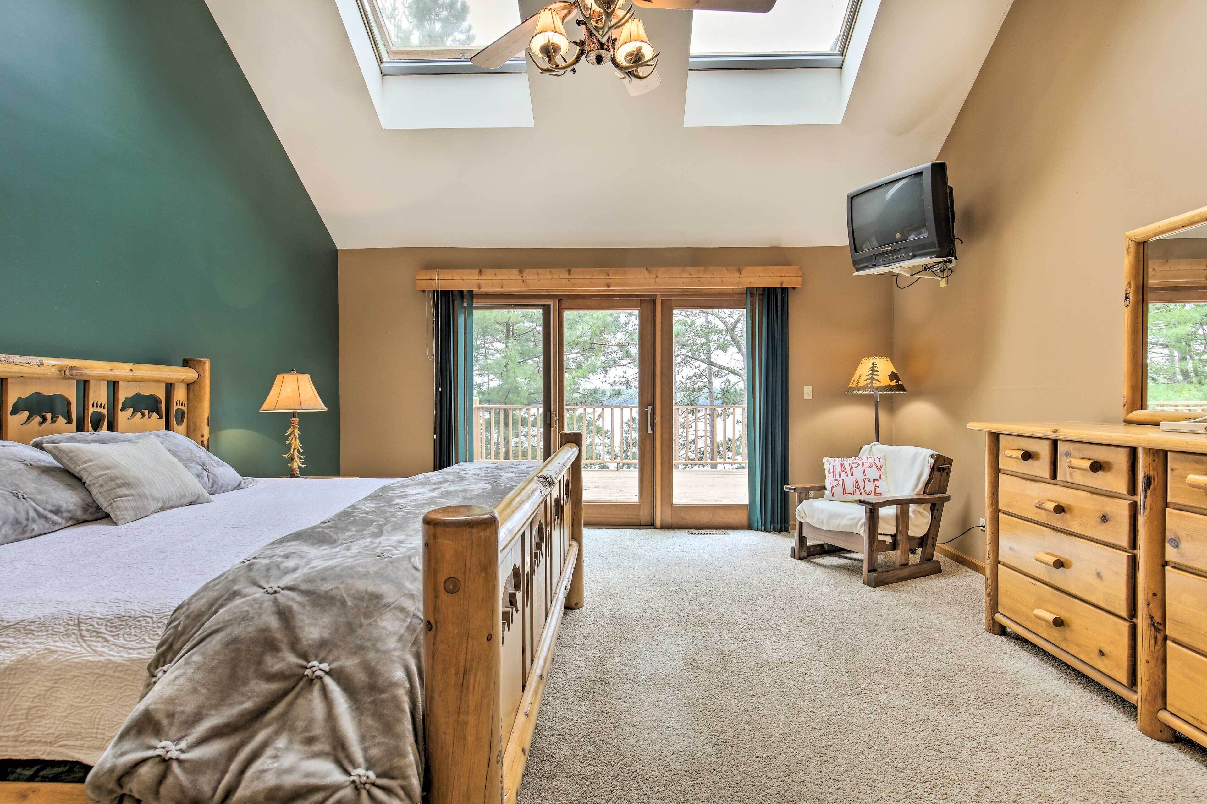 A bedroom with a bed, a wooden dresser, a TV mounted on the wall, and a balcony with a 'Happy Place' sign.
