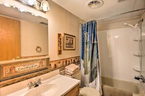 A bathroom with a sink, mirror, and shower. There is a decorative welcome sign above the sink and a shower curtain with a pattern.