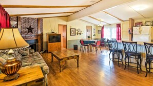 A spacious living room with a wooden floor, a sofa, a coffee table, and a TV. There is a dining area with a table and chairs, and a kitchen area with a refrigerator and a stove. - Waterfront House w/ Boat Slip on Lake Eufaula! (Canadian)