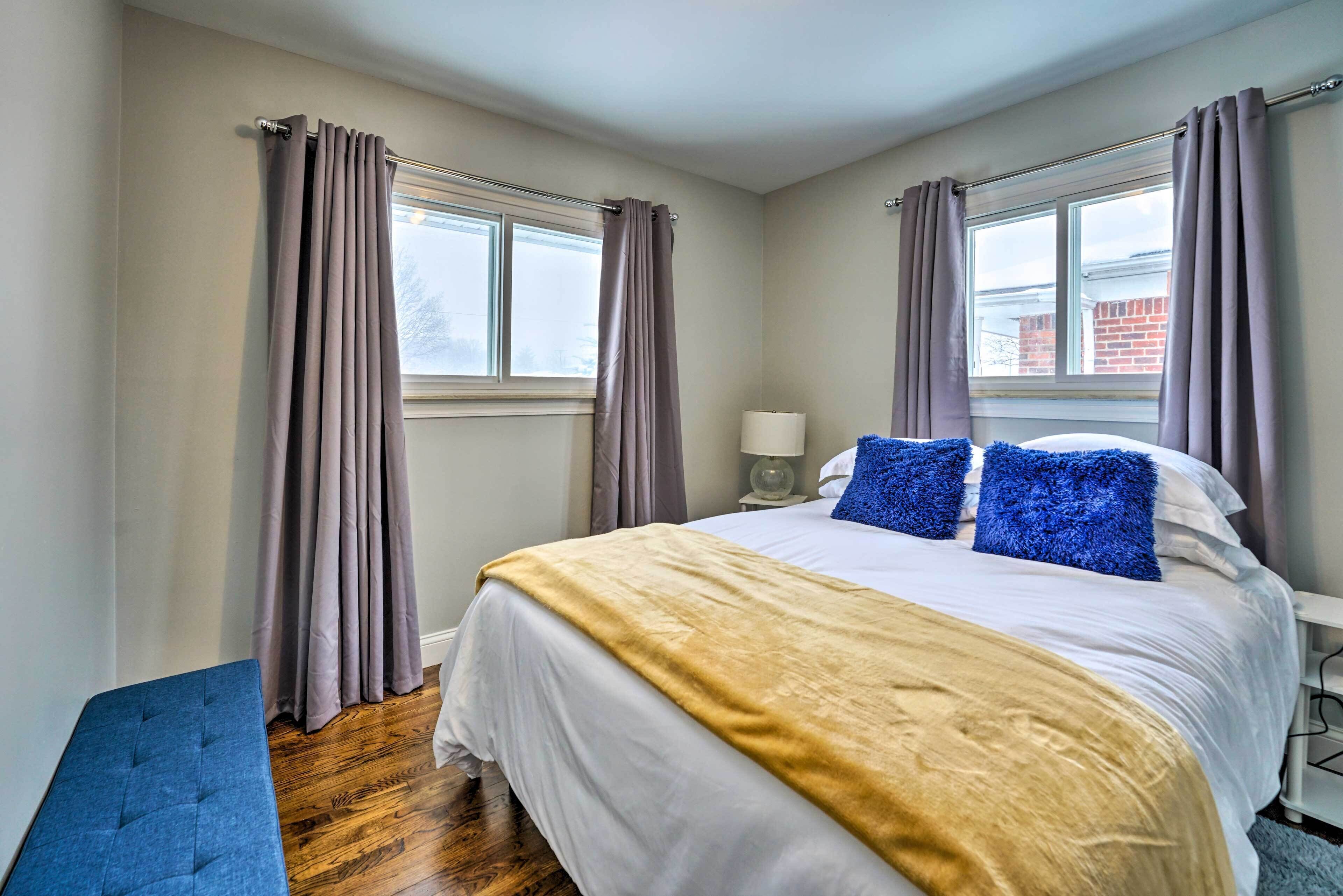 Un dormitorio con cama, un banco, dos ventanas con cortinas y una mesita de noche con lámpara.