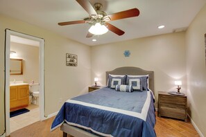 Un dormitorio con cama, mesitas de noche, ventilador de techo y un baño visible a través de una puerta abierta.
