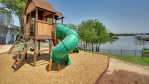 Ein Spielplatz mit einer Holzrutsche und Kletterbereich, umgeben von einem See und einem Zaun.